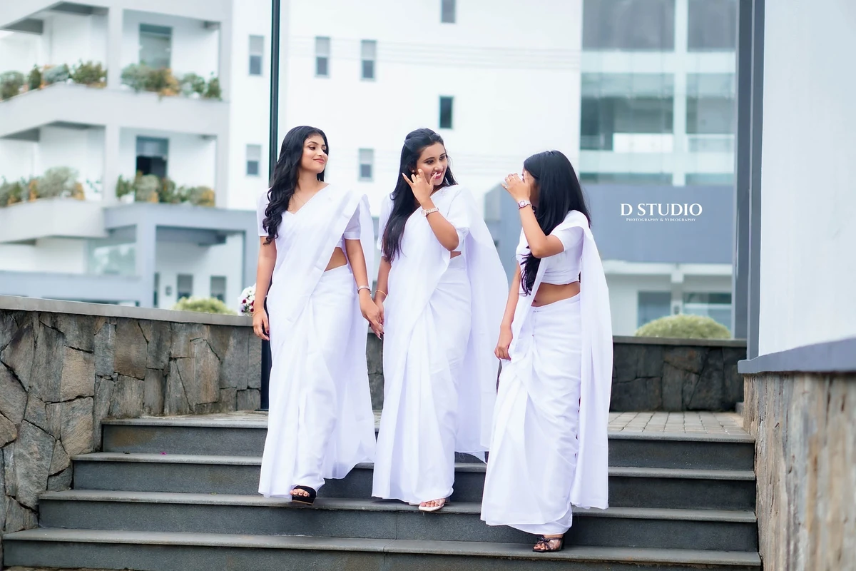 Three friends in white sarees standing together on campus steps, photographed by D Studio